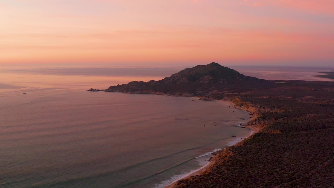 Elevated sunset view along Cabo Pulmo's coast, with the beach edge lower right, smooth water stretching left, silhouetted hills near the horizon, and no visible people.