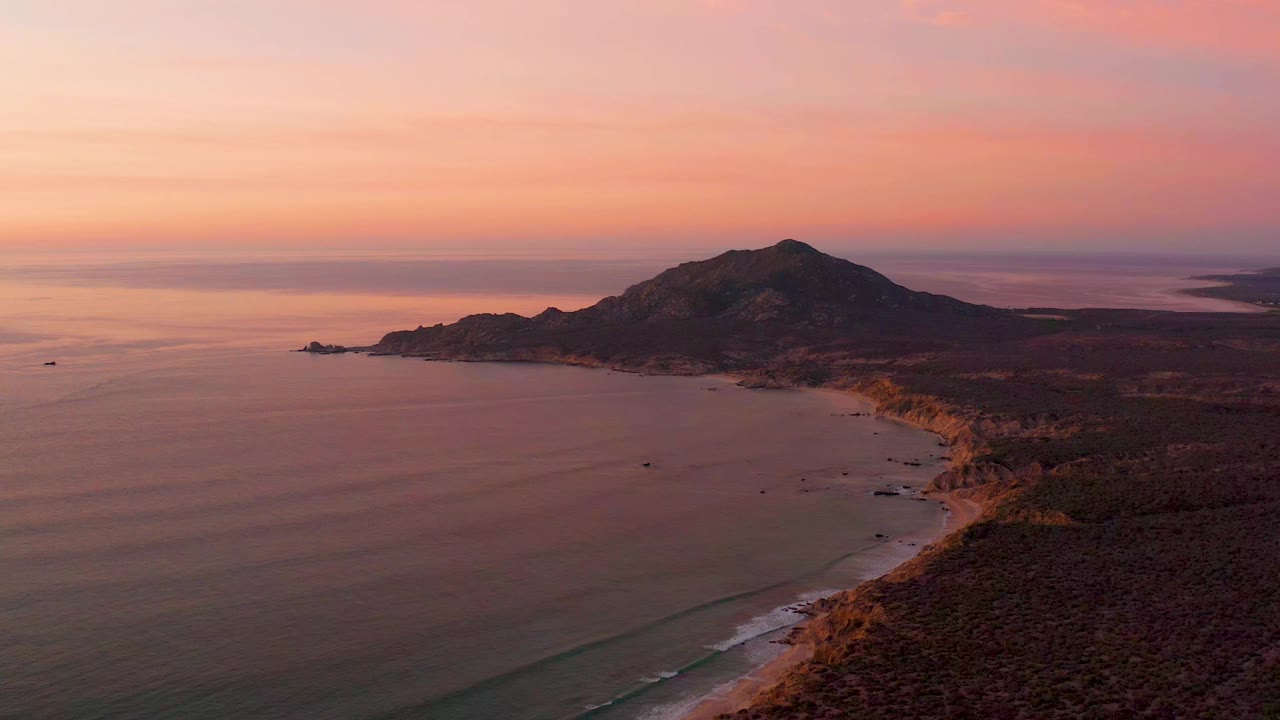 At 0:01, the Cabo Pulmo shore remains in a high wide view, with gentle wave lines near the beach, shadowed land to the right, the hill centered beyond, and no visible people.