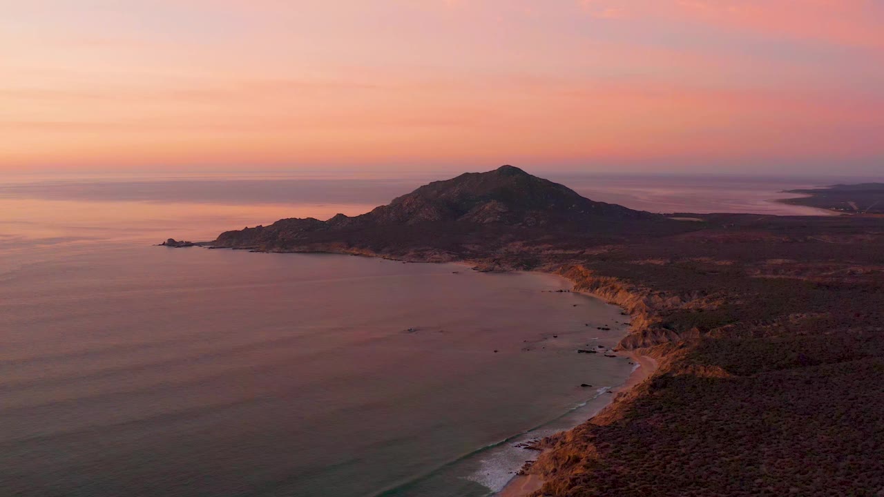 Cabo Pulmo coastline seen from above with the hill mass centered, glassy ocean to the left, a narrow beach tracing the shore, and no visible people.