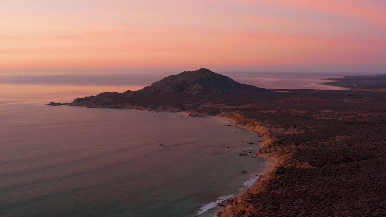 Aerial view of a bay, curving shoreline, and dark hill under a pink orange sunset sky.