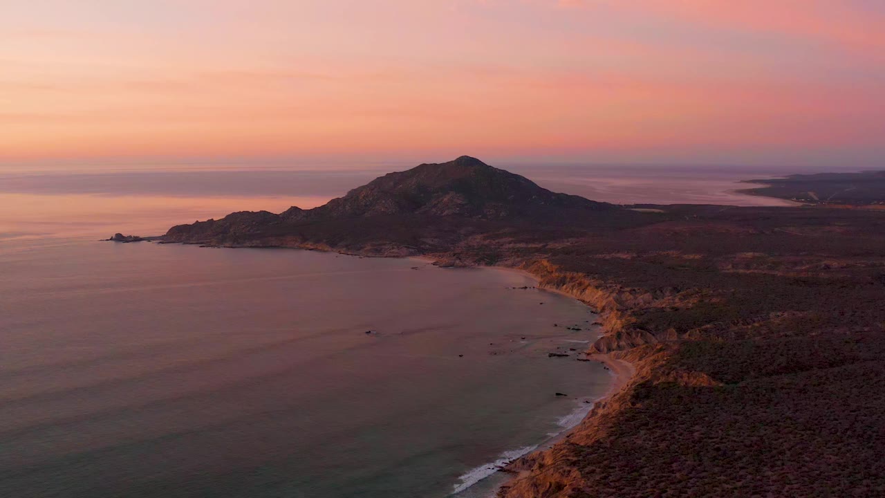 Aerial Cabo Pulmo coastline view with the ocean spread across the left side, the beach arc running into the distance, a hill centered near the horizon, and no visible people.