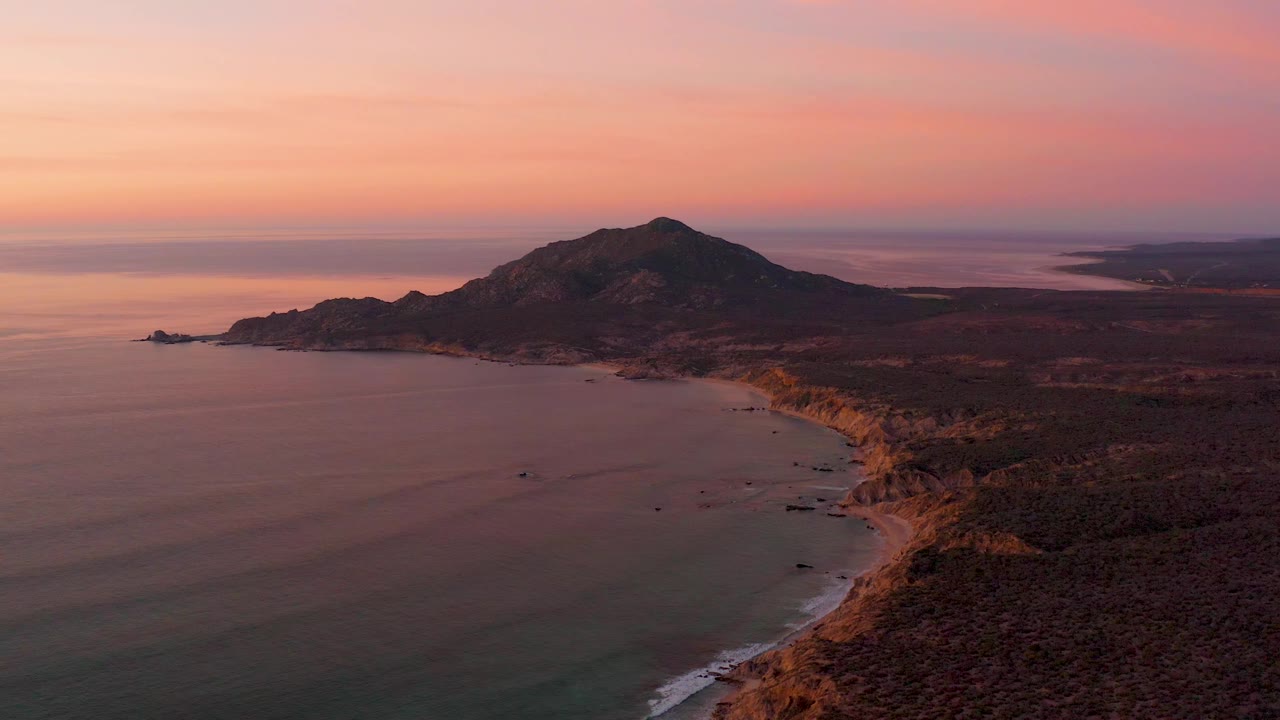 High aerial frame over Cabo Pulmo showing a broad bay at left, a shadowed shoreline and beach at right, the hill set farther back, soft pink orange sky, and no visible people.