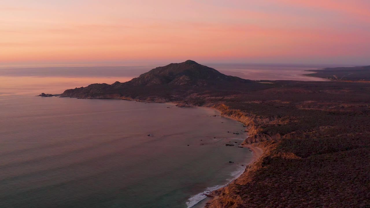 Mid-clip aerial view of Cabo Pulmo with darker foreground coast, calm water to the left, a warm-lit shoreline bend, hill mass in the background, and no visible people.