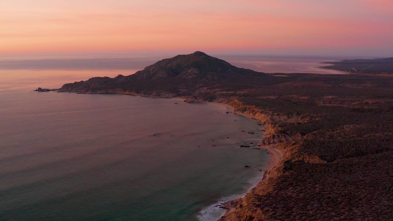 Aerial sunset view over Cabo Pulmo coastline with a dark mountain behind a rocky shore, calm ocean in the foreground, and no visible people.