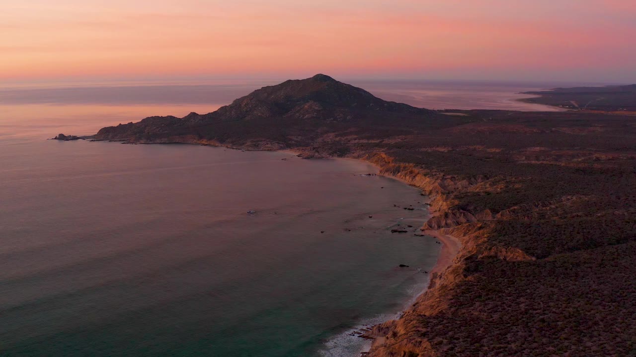 Cabo Pulmo aerial frame with the shoreline running diagonally below a dark mountain, orange light on the cliffs, calm water, and no visible people.