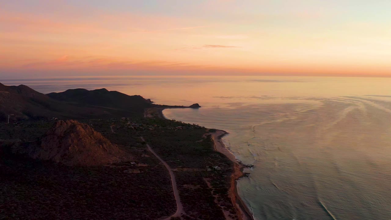 Elevated Cabo Pulmo coastline frame with the rocky hill more central, the road running toward the beach, calm sea to the right, and no visible people.