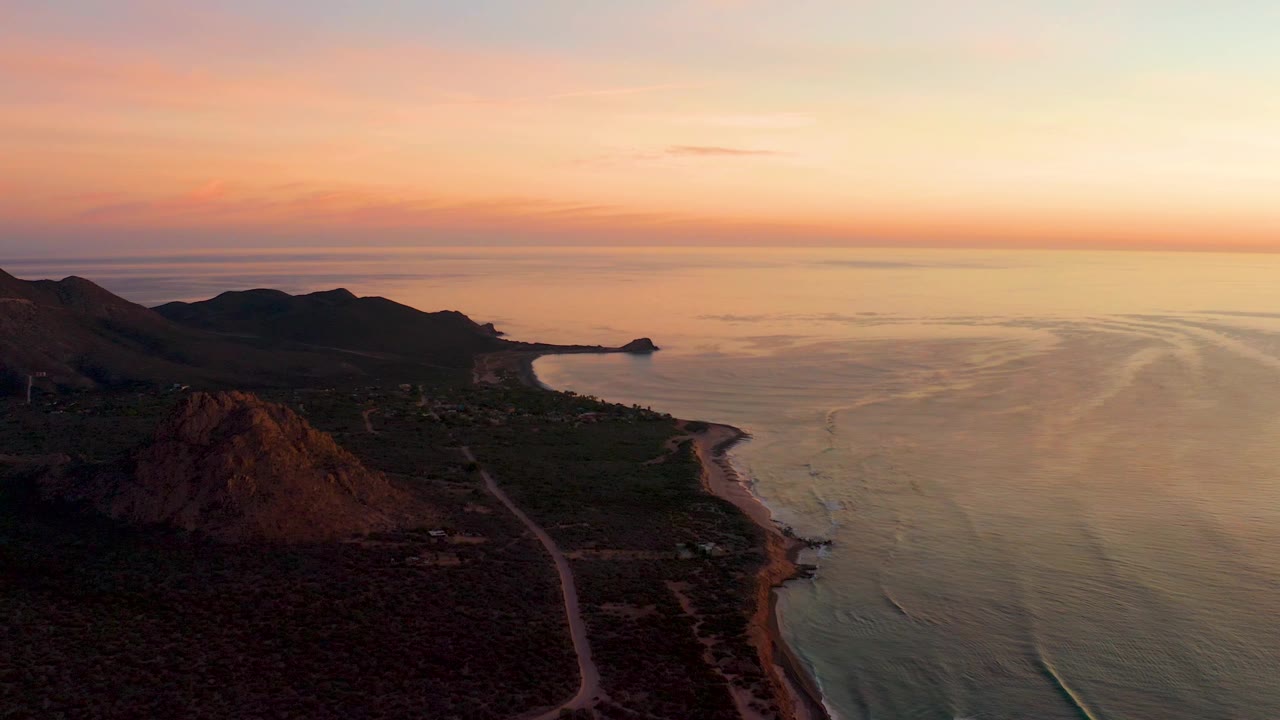 Cabo Pulmo aerial frame showing mountain silhouettes on the left, beach and shoreline below, reflective ocean bands on the right, and no visible people.