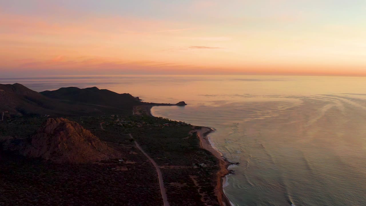 At 0:01, Cabo Pulmo coastline fills the frame with a shadowed hill in the lower left, a straight road near the beach, open water to the right, and no visible people.