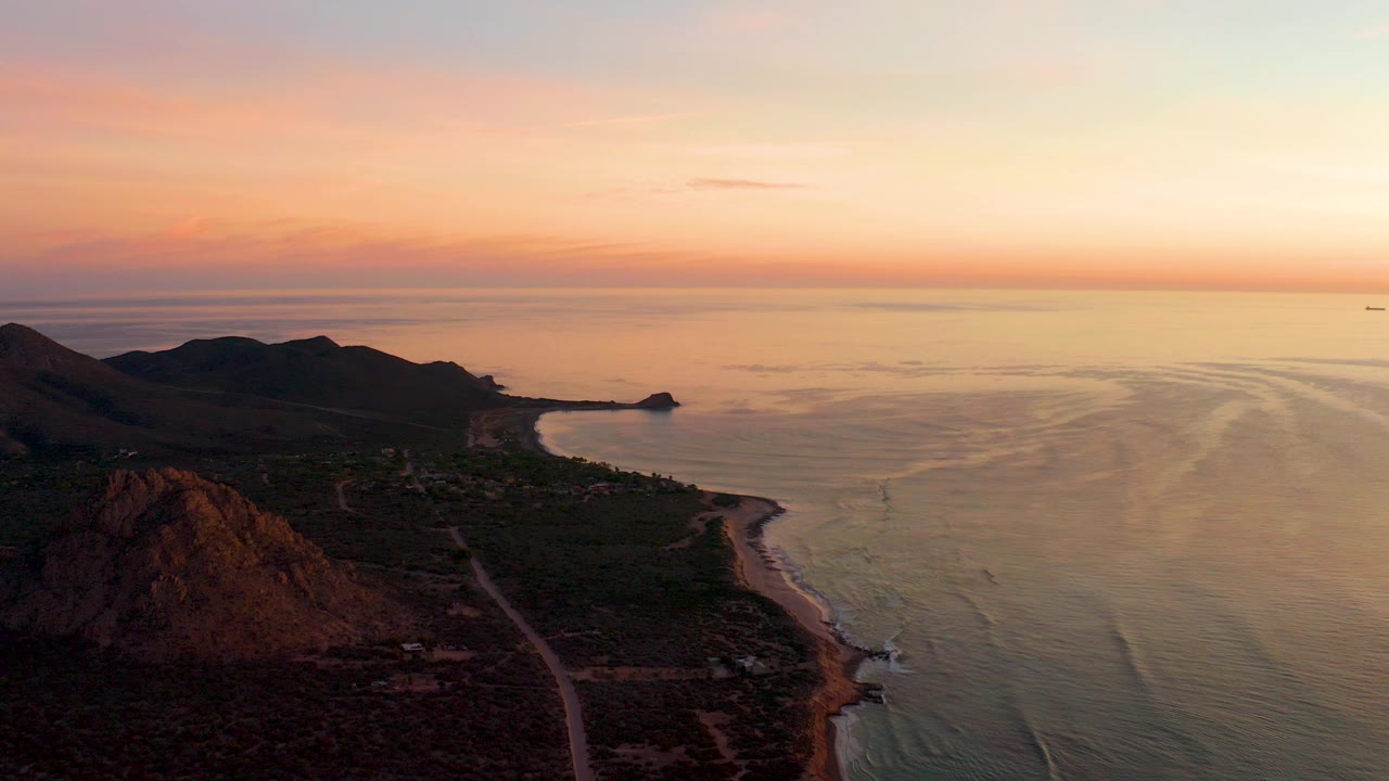 At 0:00, an elevated view over Cabo Pulmo shows dark hills and scrubland beside a shoreline road, with warm sunset color over calm ocean water and no visible people.
