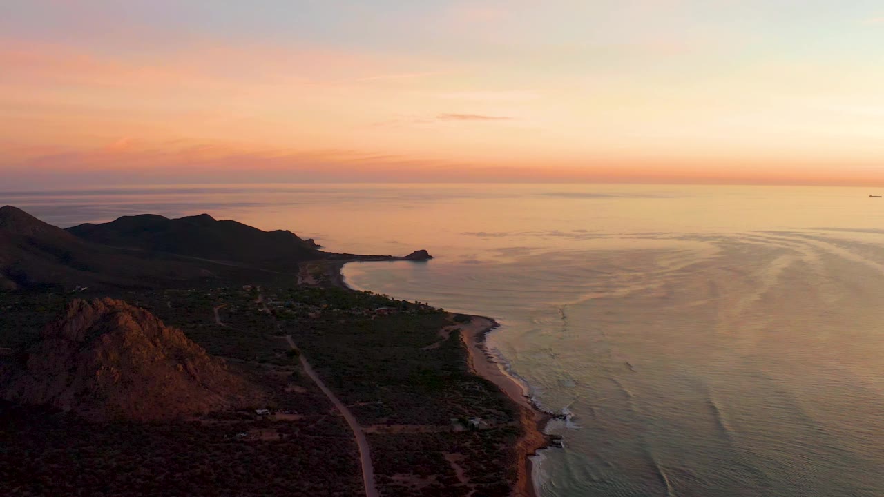 At 0:01, the Cabo Pulmo coastline remains in a wide aerial view, with the beach curve leading toward a small rocky point, rippled ocean at right, and no visible people.