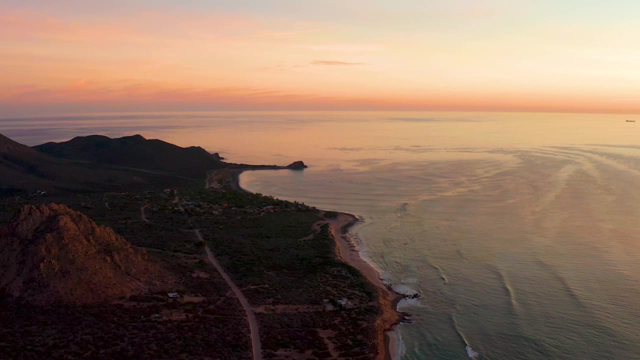 Cabo Pulmo coastline from above with the mountain ridge dark against the sunset sky, the road running near the shore, small wave bands in the water, and no visible people.