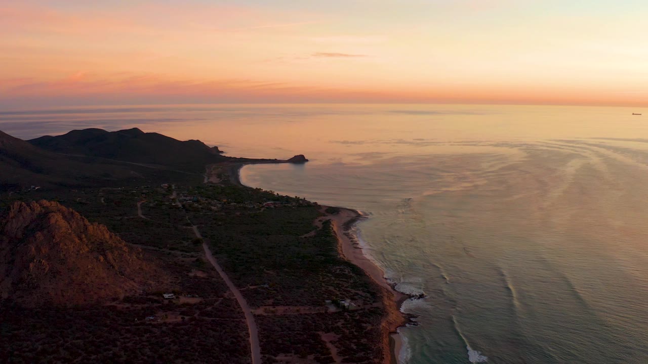 Aerial sunset view in Cabo Pulmo with the shore road more centered below the mountains, low surf tracing the beach edge, warm sky over the horizon, and no visible people.