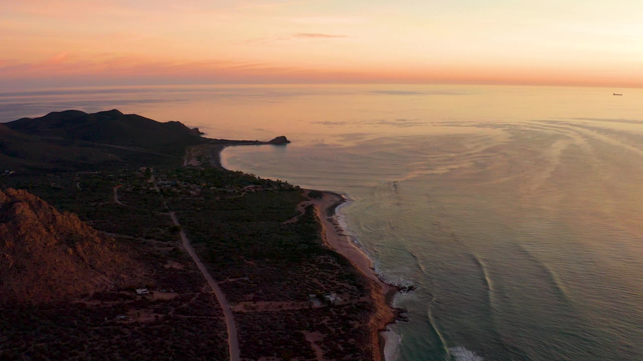 Aerial sunset view over Cabo Pulmo with a winding coastal road below, a narrow beach along the right edge of land, dark hills in the distance, orange sky, and no visible people.