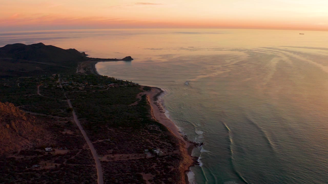 Aerial view over Cabo Pulmo at sunset with a road along scrub-covered coast, beach and hills at left, ocean wave lines at right, a small distant boat near the horizon, and no visible people.