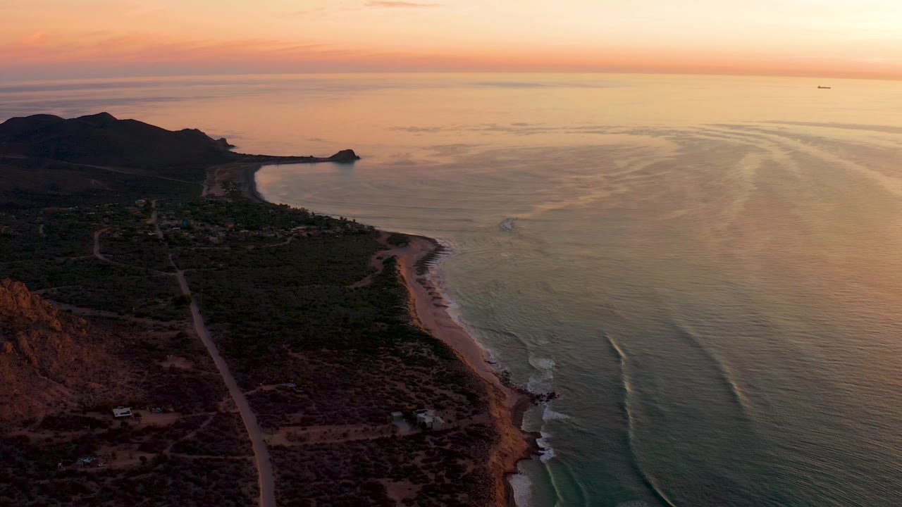 High aerial frame over Cabo Pulmo showing the beach line slightly farther right in the composition, dark hillside foreground, pale wave streaks on the ocean, and no visible people.