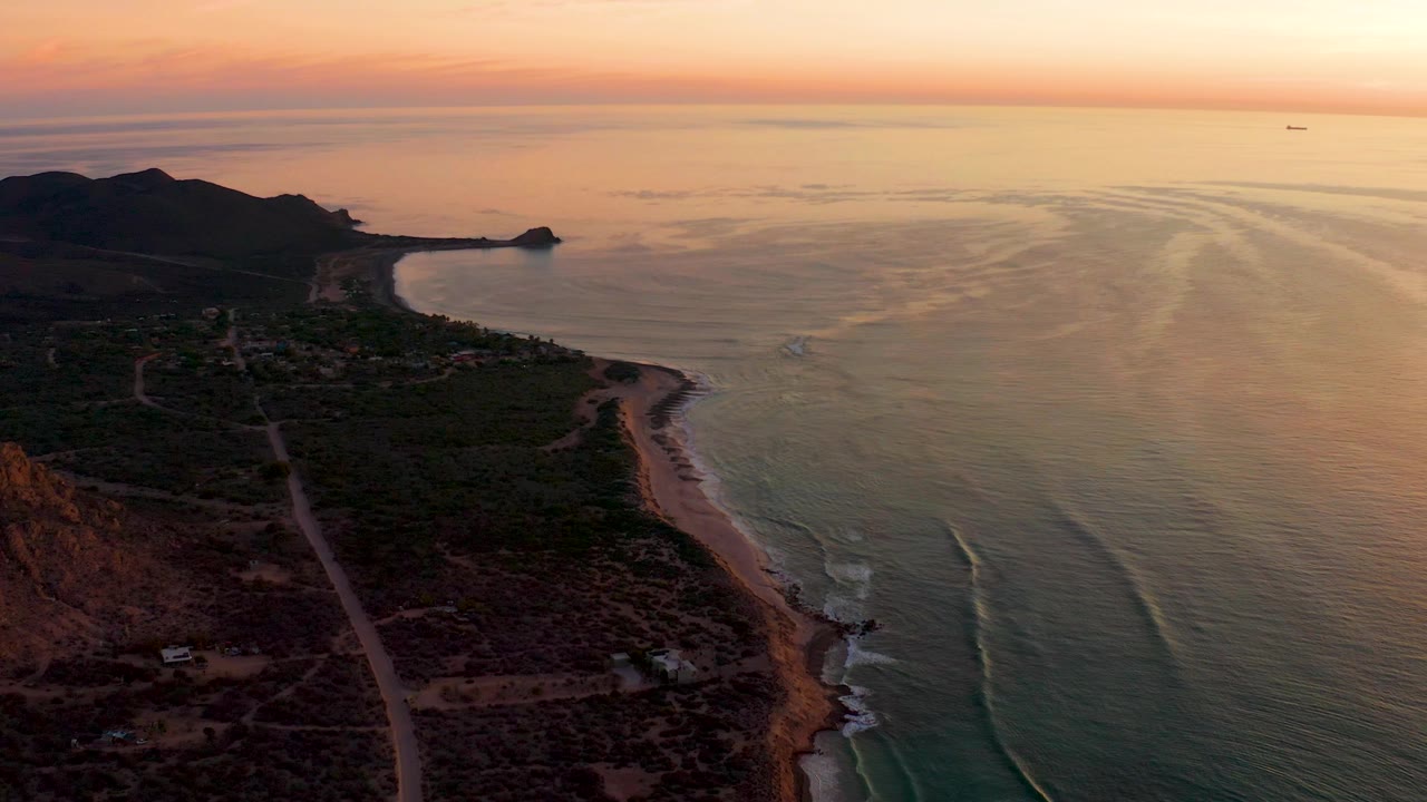 Sunset aerial with dark hills, coastal vegetation, beach, and calm ocean.