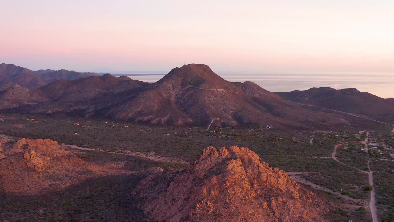 Wide aerial view over Cabo Pulmo desert mountains with reddish foreground rock, a valley road, layered ridges, distant water, pink-orange sky, and no visible people.