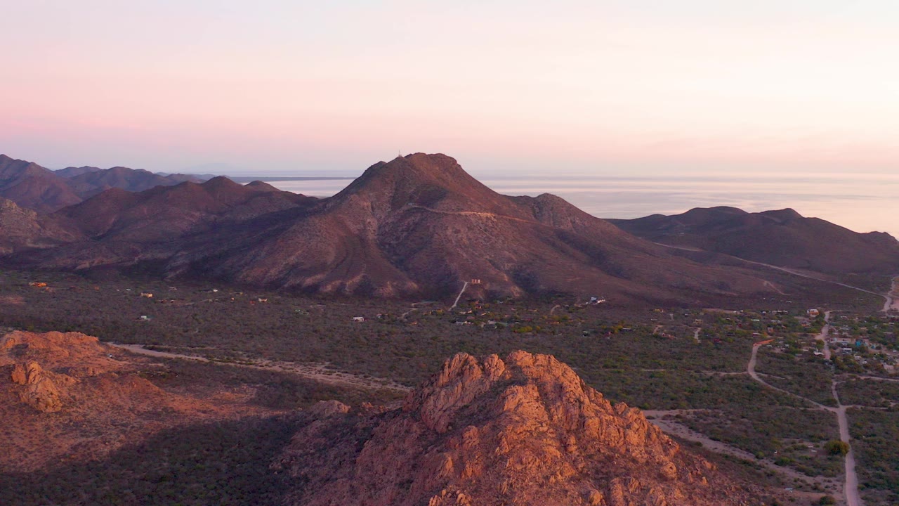 Wide aerial view over Cabo Pulmo desert terrain with reddish foreground rock, a central mountain ridge, faint dirt roads in the valley, distant water on the horizon, and no visible people.