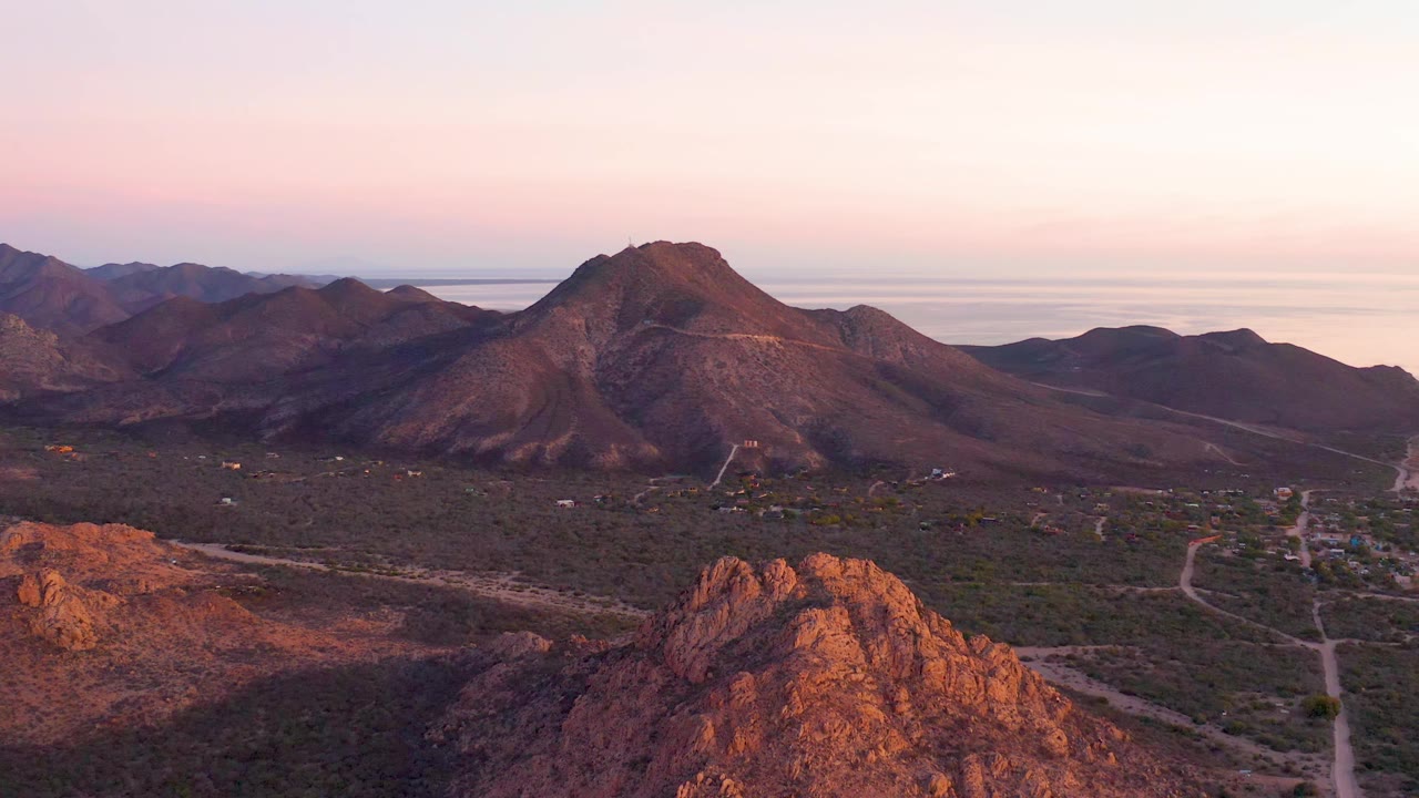 Cabo Pulmo aerial frame showing a broad arid valley below a dark mountain ridge, reddish rocks in the lower foreground, pale water beyond the hills, and no visible people.