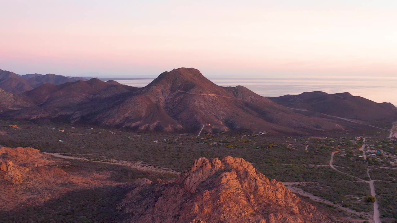 A wide Cabo Pulmo dusk view frames layered mountains across the background, a scrub-covered valley with dirt tracks, reddish rock at the bottom edge, and no visible people.