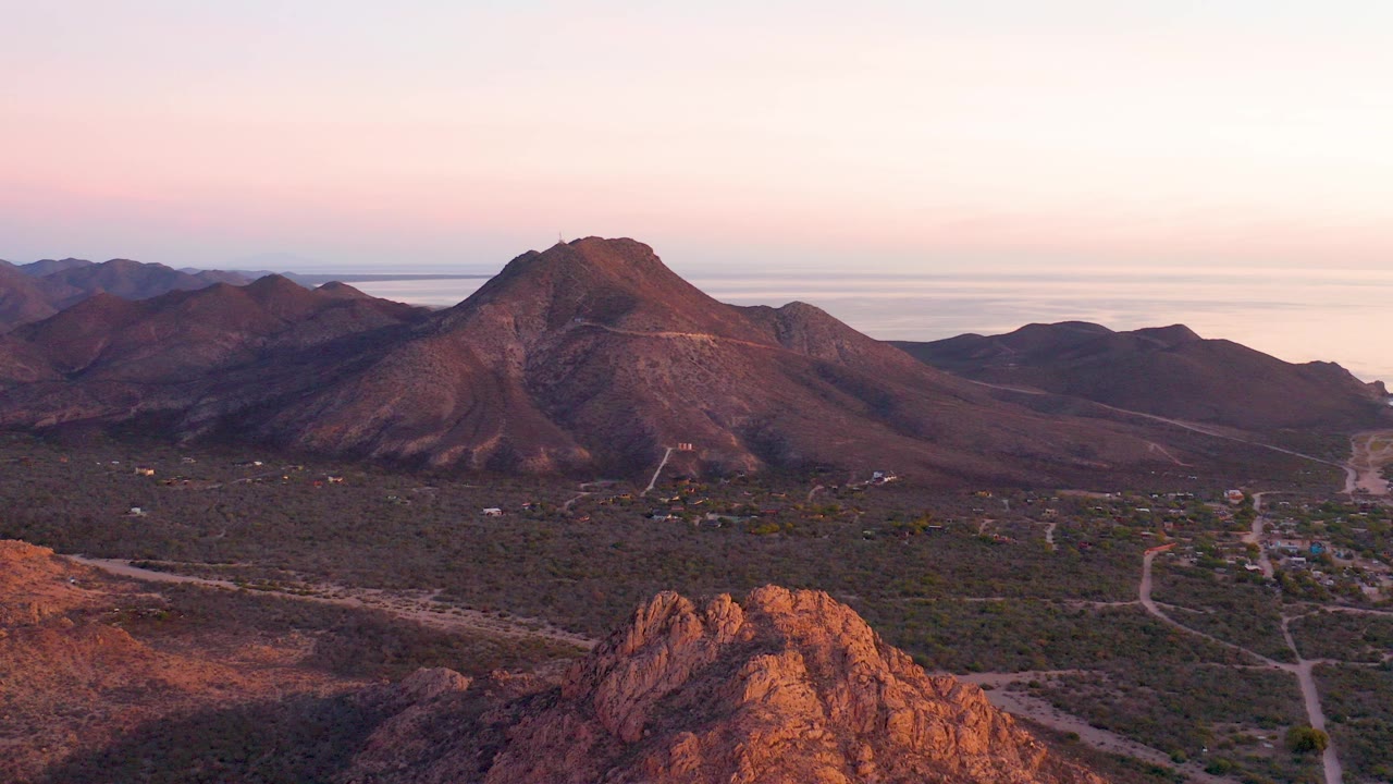 Aerial view in Cabo Pulmo, Baja California Sur showing a sunlit rocky foreground ridge, scrub valley roads, dark mountains, distant coastal water, pink dusk sky, and no visible people.