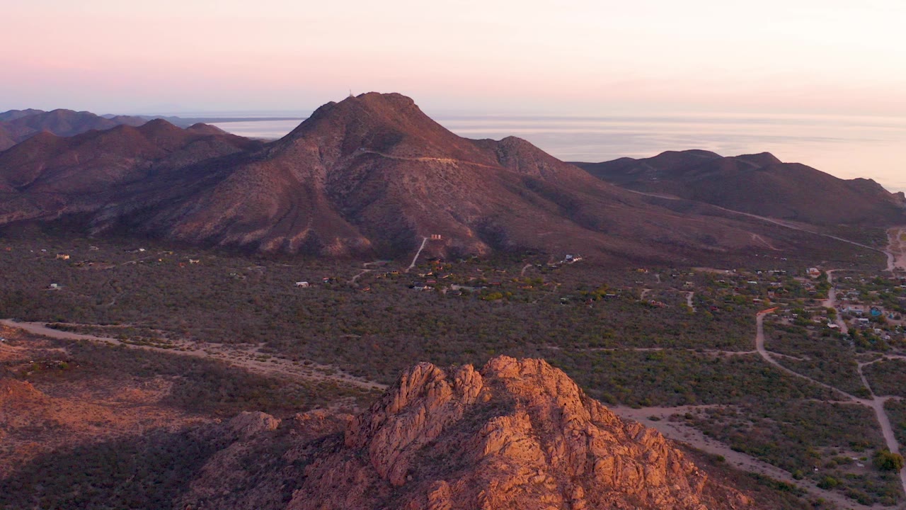 Aerial view in Cabo Pulmo, Baja California Sur, showing a foreground rock outcrop, desert valley roads, distant mountains, a pale sea horizon, and no visible people.
