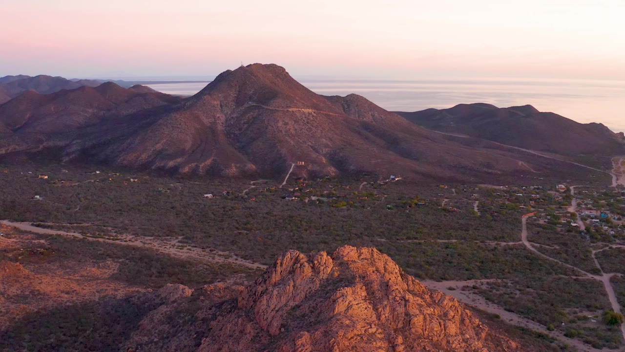 Cabo Pulmo aerial frame with the central mountain ridge slightly more centered, orange foreground rocks, sparse desert vegetation, dirt roads, distant water, and no visible people.