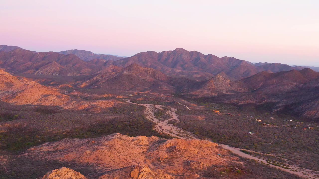 High view over Cabo Pulmo scrubland where the dirt road bends through the center of the valley below sunlit orange hills and darker mountain ridges, with no visible people.
