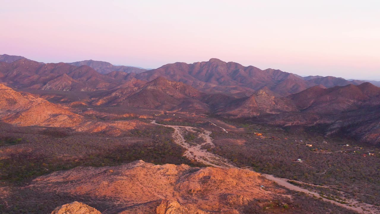 Aerial composition over Cabo Pulmo shows warm light hitting the left hillside, a pale road running through the valley floor, distant peaks, and no visible people.