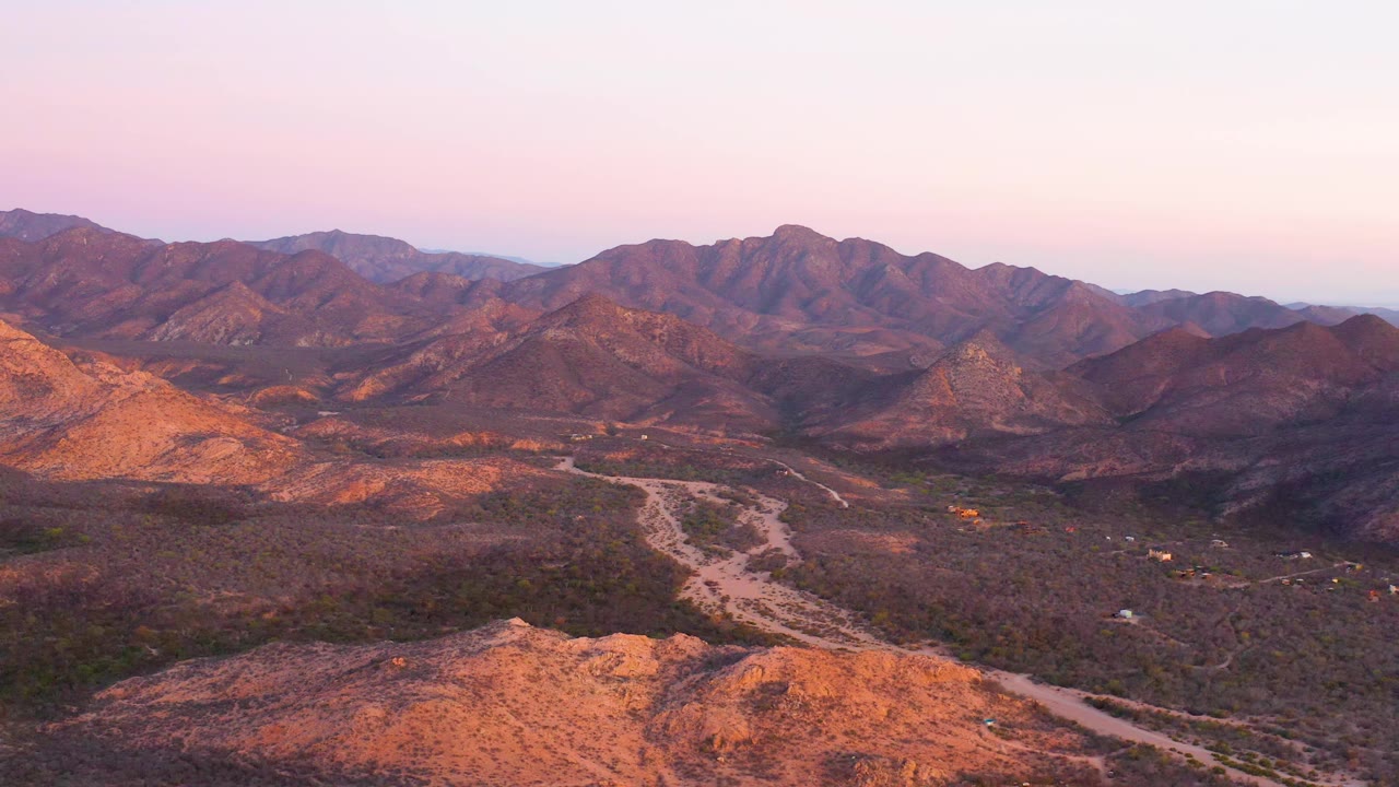Wide aerial frame in Cabo Pulmo showing the pale road bending through scrub terrain, reddish ridges in the background, warm side light, and no visible people.