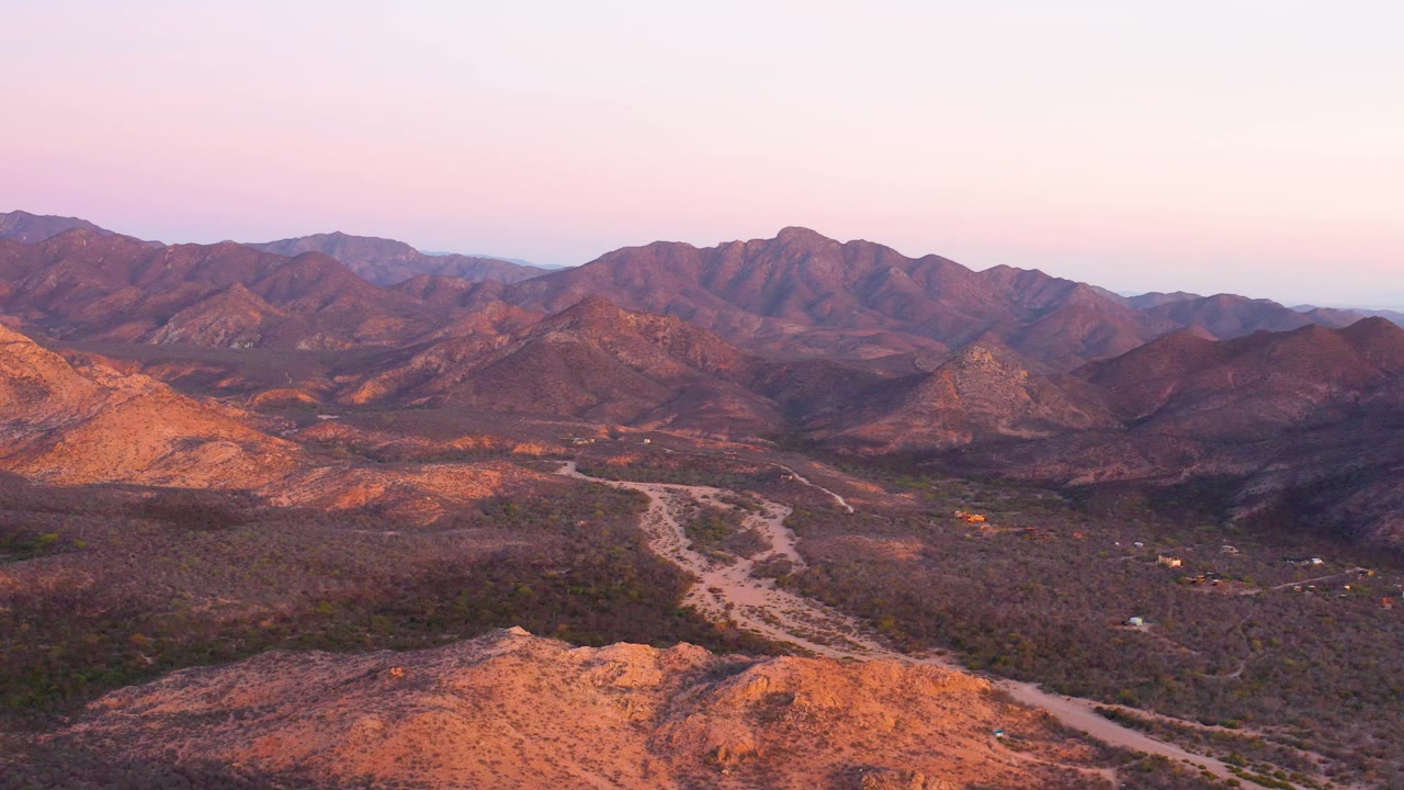 Cabo Pulmo desert panorama with a winding road running across the valley floor, orange-lit foreground hills, darker scrub patches, distant ridges, and no visible people.