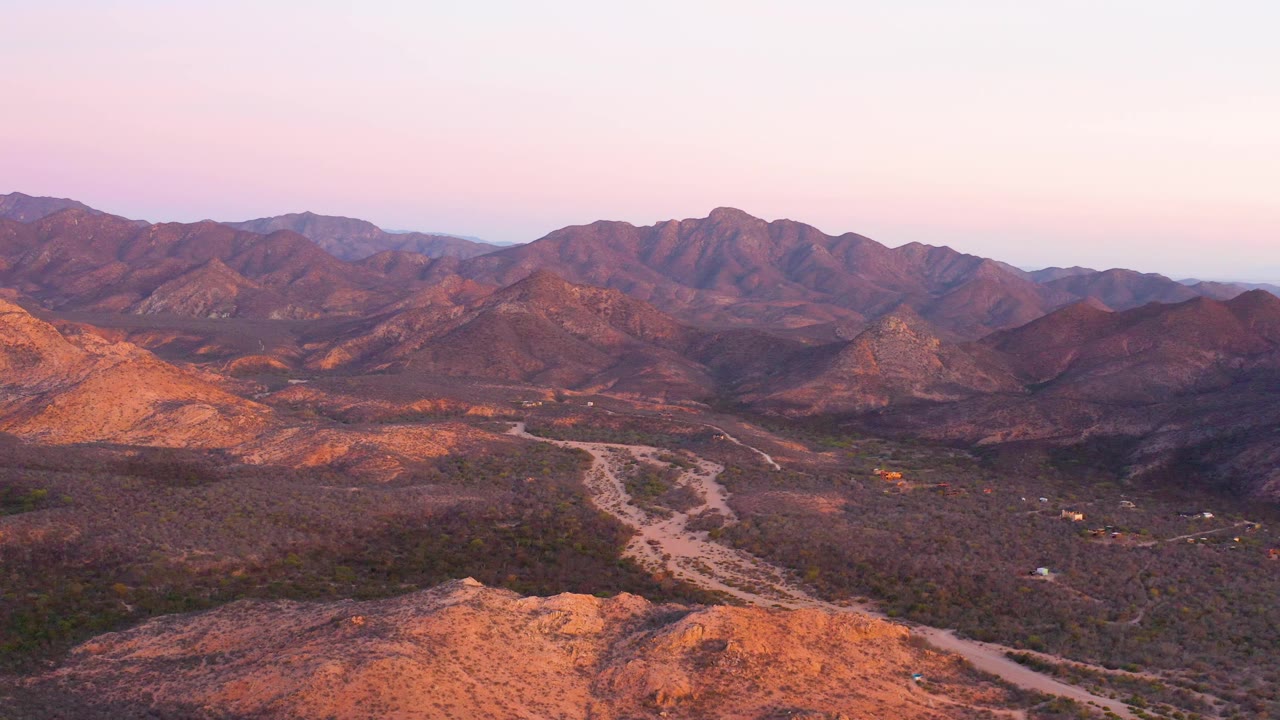 Aerial Cabo Pulmo view with the road threading through low desert vegetation, mountain ridges filling the horizon, warm orange highlights on slopes, and no visible people.