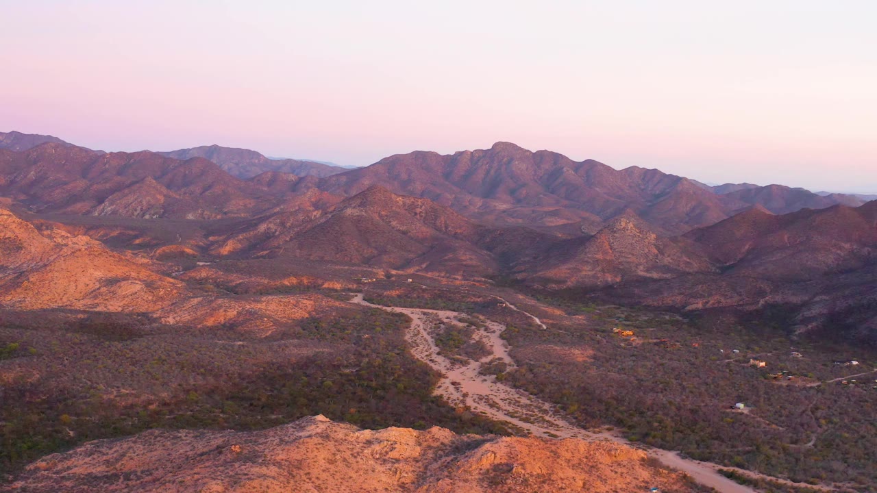 One second in, the Cabo Pulmo aerial view holds on layered brown mountains, a dry valley floor, a pale wash running through the midground, soft pink sky, and no visible people.