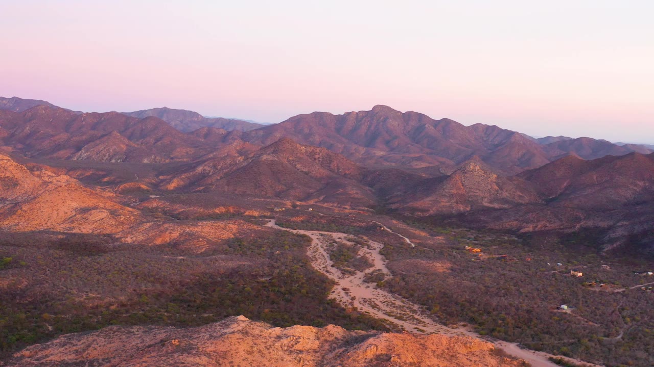 At 0:03, the Cabo Pulmo valley remains in a high wide composition, with the pale channel and road lower in frame, rugged mountains across the horizon, soft pink sky, and no visible people.