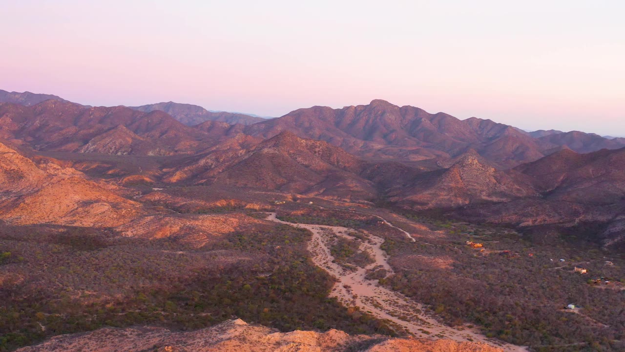 Wide aerial frame in Cabo Pulmo showing layered rocky hills, scrubby desert floor, a pale branching wash or road through the valley, soft pink-orange sky, and no visible people.