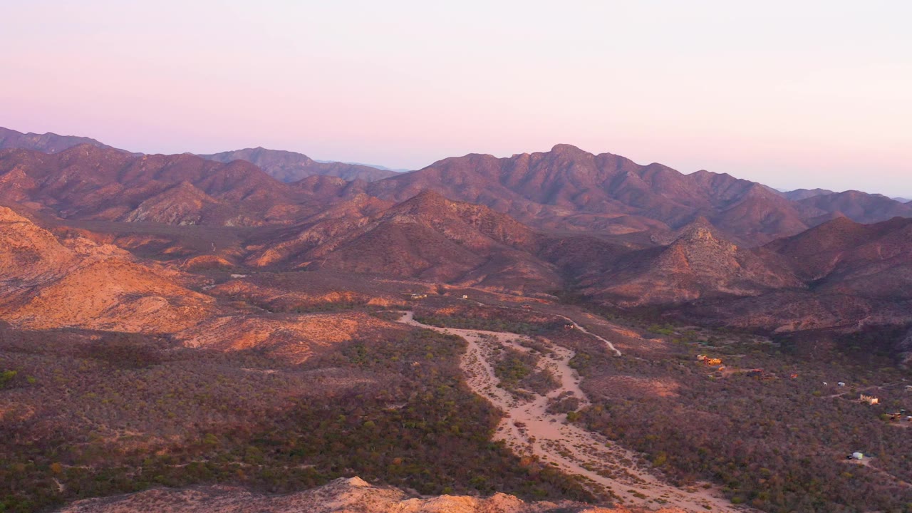 Elevated view across Cabo Pulmo desert mountains with a pale wash or road curving through scrubland, warm highlights on rocky slopes, pink evening sky, and no visible people.