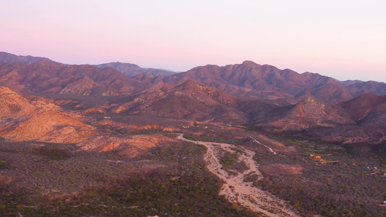 Wide Cabo Pulmo aerial frame with the dirt road centered lower in the valley, brown ridges across the horizon, warm light on the left hillside, and no visible people.