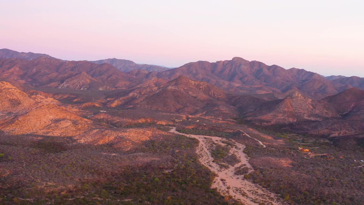 Aerial frame over Cabo Pulmo terrain where the pale road tracks through a scrub valley below rugged hills, with sunrise color on the slopes and no visible people.