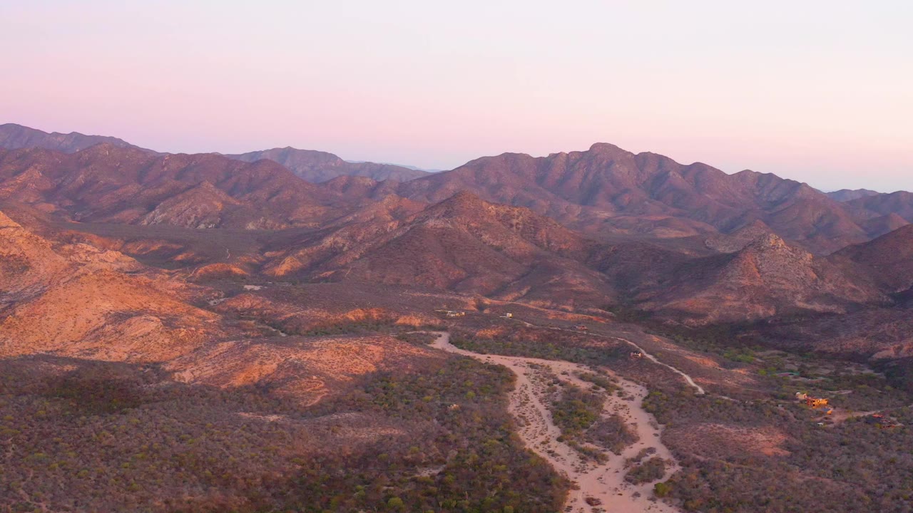 Aerial view over Cabo Pulmo desert scrub and a pale dirt road leading toward layered mountains under a pink dusk sky, with no visible people.