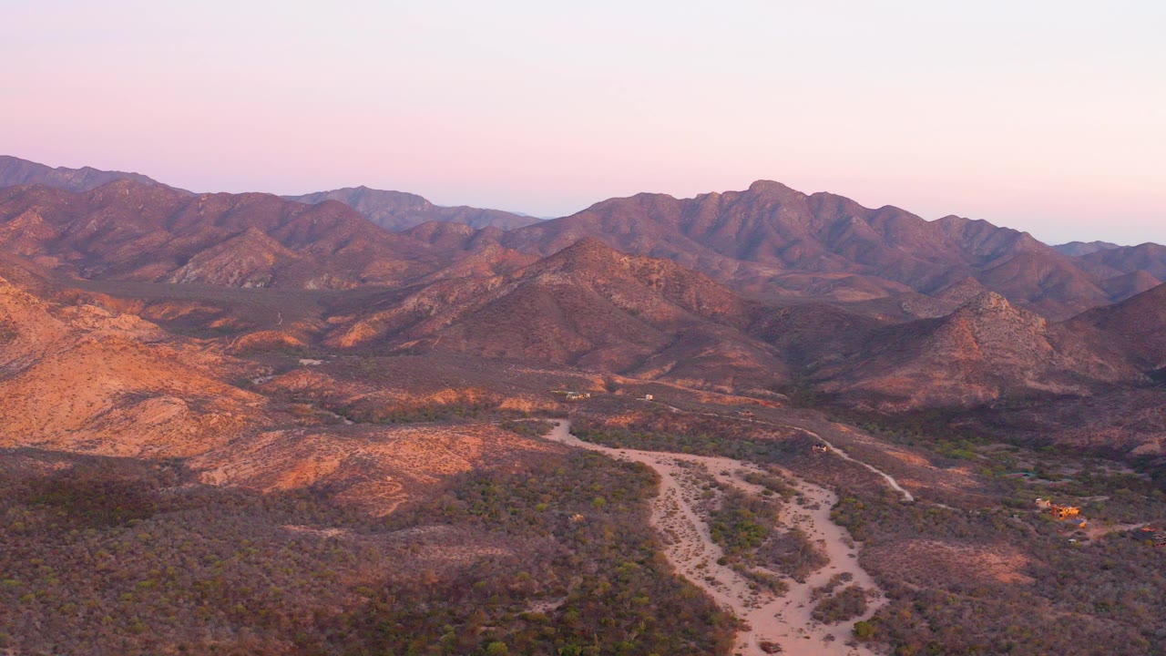 Aerial view in Cabo Pulmo, Baja California Sur, showing a pale dirt road crossing scrub desert toward layered mountains under a pink dusk sky, with no visible people.