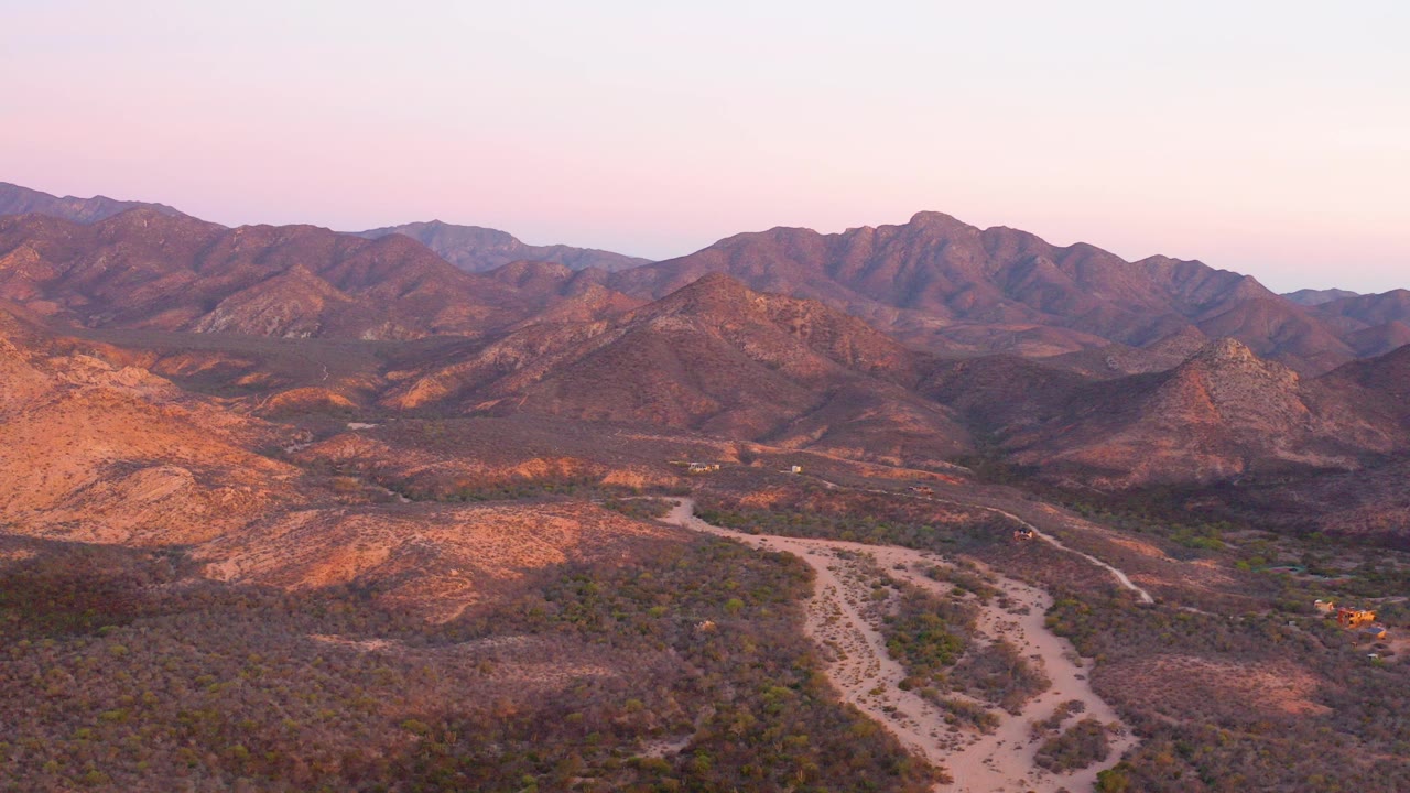 Opening frame of Cabo Pulmo, Baja California Sur desert mountains with a pale dirt road splitting the scrub valley below a pink-blue sky, no visible people.
