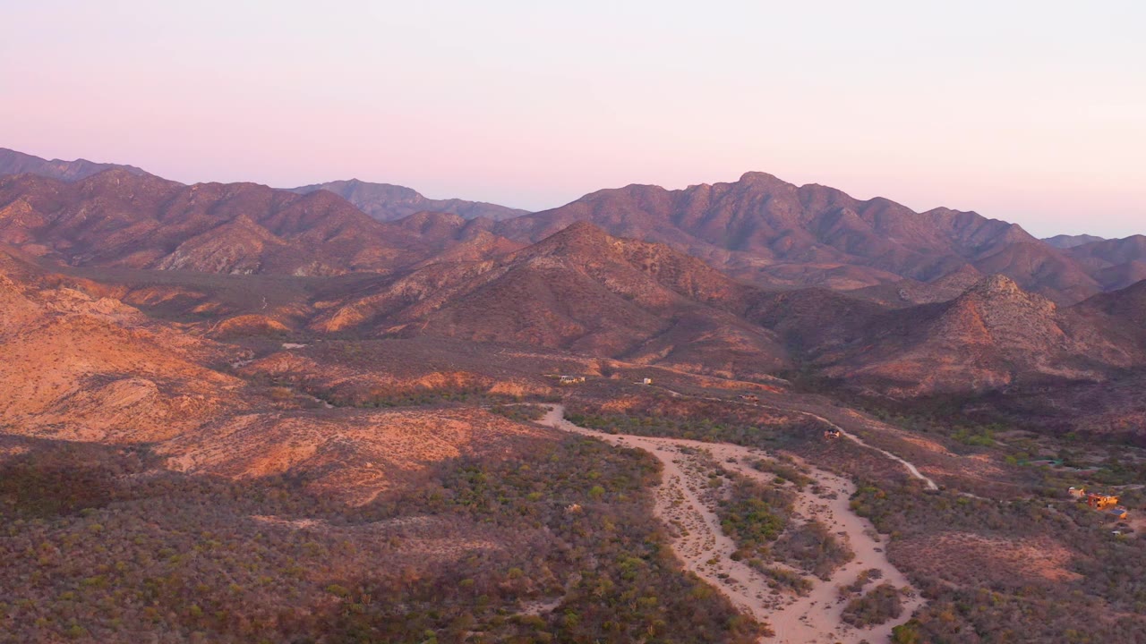 Early frame showing the winding Cabo Pulmo dirt road more centered in the valley, with orange-lit hills and distant Baja California Sur ridges, no visible people.