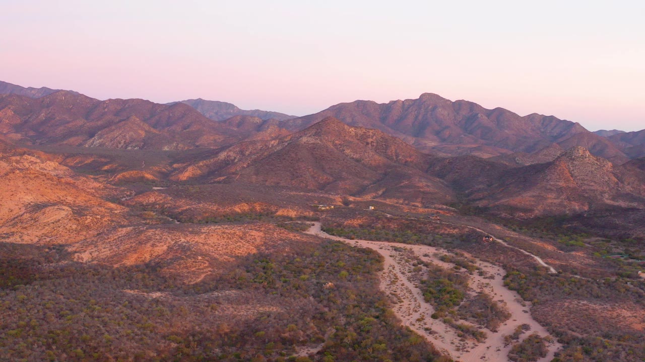 Aerial view over Cabo Pulmo desert terrain with a pale dirt road crossing scrubby hills toward layered mountains under a pink dusk sky, no visible people.