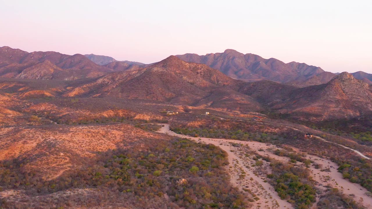 Aerial frame over Cabo Pulmo, Baja California Sur, Mexico, with dry hills, sparse vegetation, a pale dirt road in the foreground, distant ridges, pink sunset sky, and no visible people.
