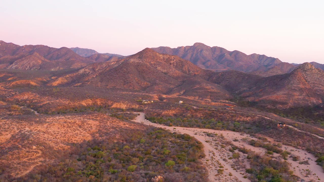 Aerial frame near Cabo Pulmo showing the dirt road bending through dry brush, with sunlit hills and layered mountains in the background, no visible people.