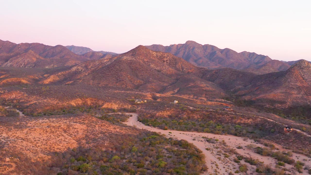 A dirt track bends through arid Cabo Pulmo terrain with low shrubs in the foreground and warm brown mountain ridges across the horizon, with no visible people.