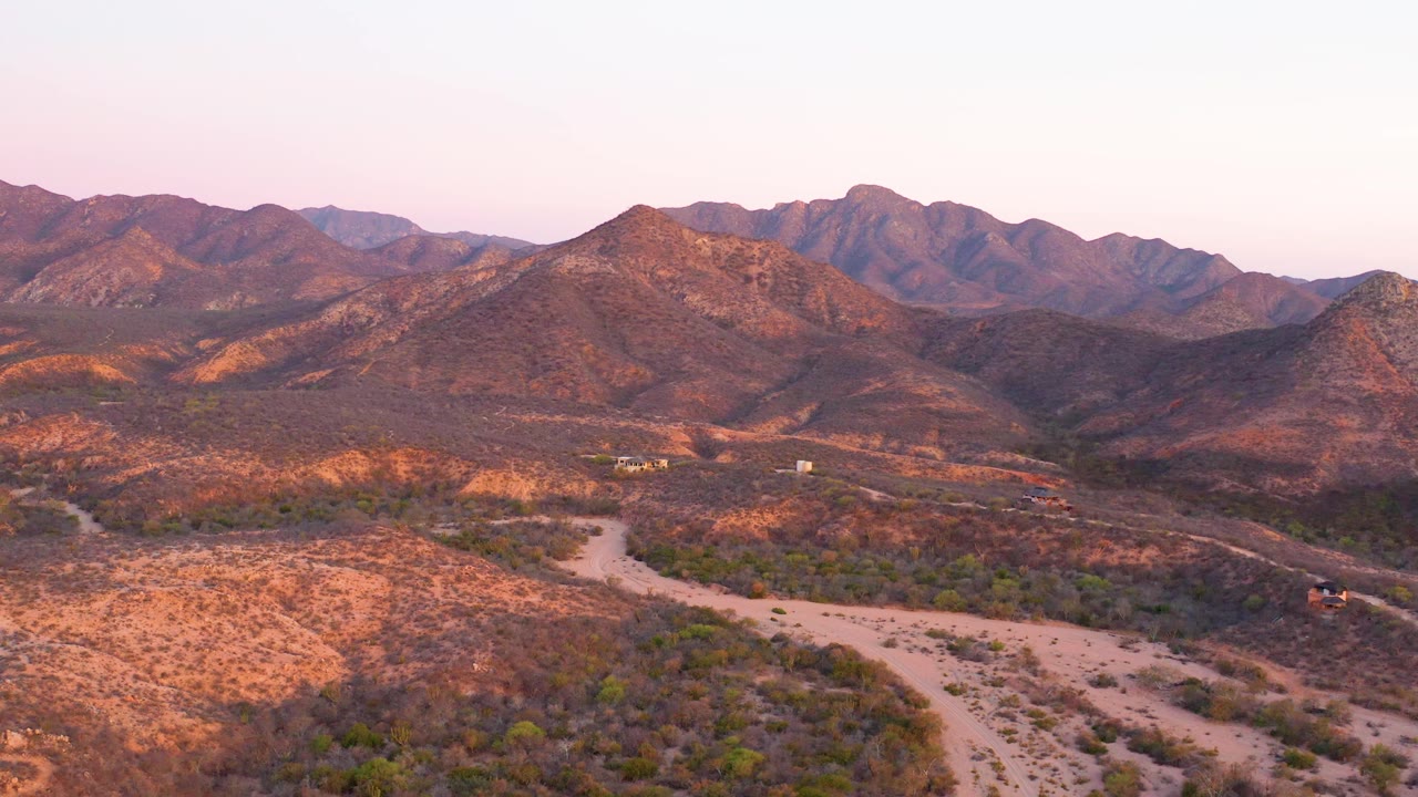 The frame shows a pale road cutting through a Cabo Pulmo desert valley, scattered vegetation, and a distant mountain range in warm dusk light, with no visible people.
