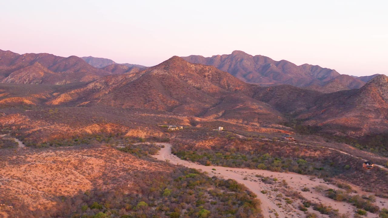 A broad Cabo Pulmo desert landscape fills the frame, with the dirt road running through scrubland and mountains layered behind it under pink evening light, with no visible people.