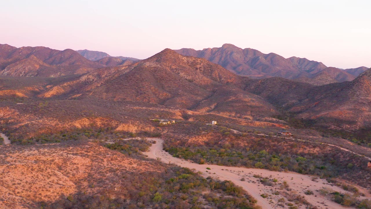 Aerial frame from Cabo Pulmo, Baja California Sur showing a pale winding dirt road across dry scrub, reddish hills, layered mountains, warm dusk light, and no visible people.
