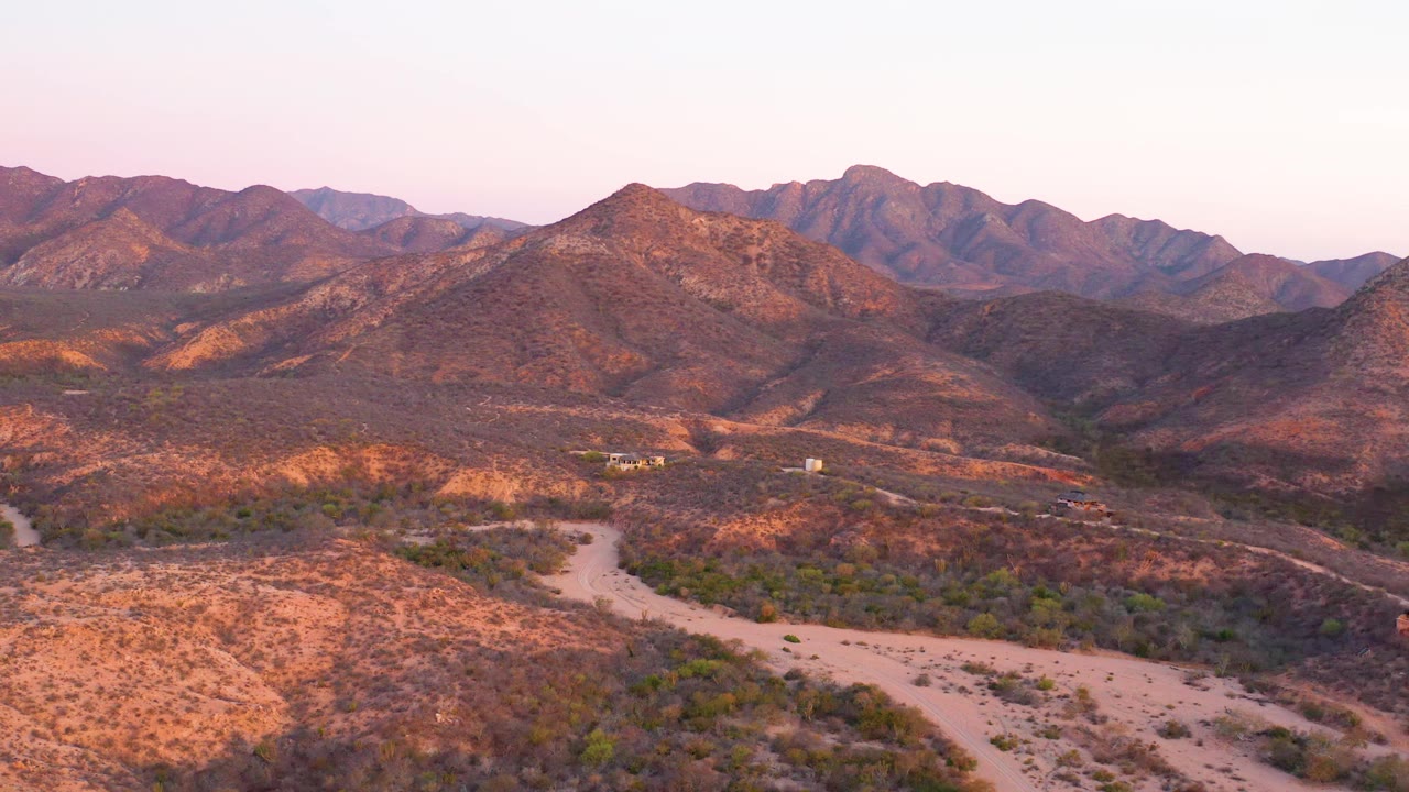Cabo Pulmo aerial view with the dirt road curving through the lower frame, scattered desert vegetation, sunlit reddish hills, distant mountains, pale sky, and no visible people.